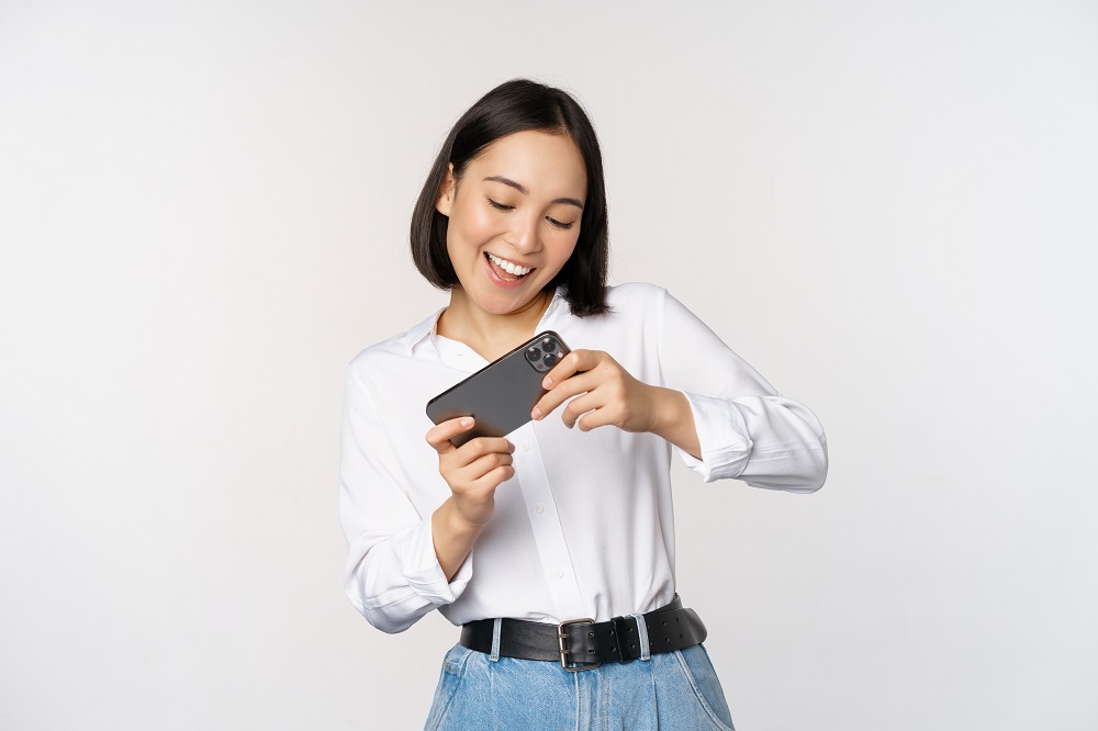 Woman playing mobile video game on smartphone, looking at horizontal phone screen, standing over white background.